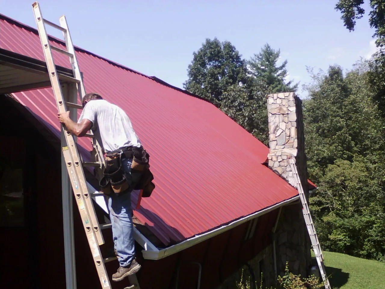 Roofer on a ladder beside a red metal roof and stone chimney — Soffit & Fascia Repair by Roofing Brothers in Brogan